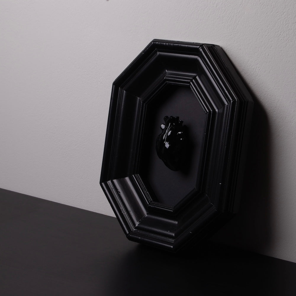 quadro con cornice ottagonale e cuore anatomico nero poggiato al muro sopra un ripiano nero