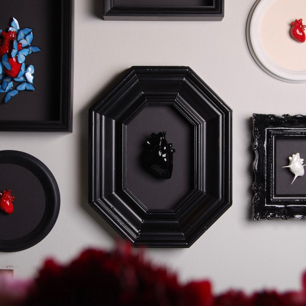 quadro con cornice ottagonale e cuore anatomico nero appeso in parete assieme ad altri quadretti con cuori di varie forme
