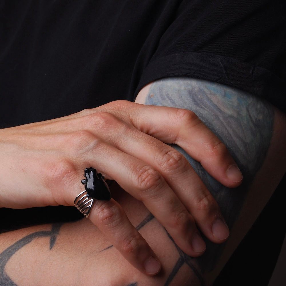 anello con cuore anatomico nero, indossato su mano maschile poggiata su braccio con tatuaggi