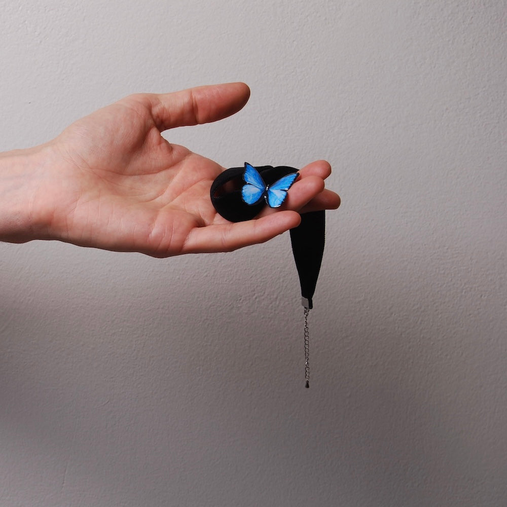 mano che tiene un choker in velluto nero con pendente farfalla blu