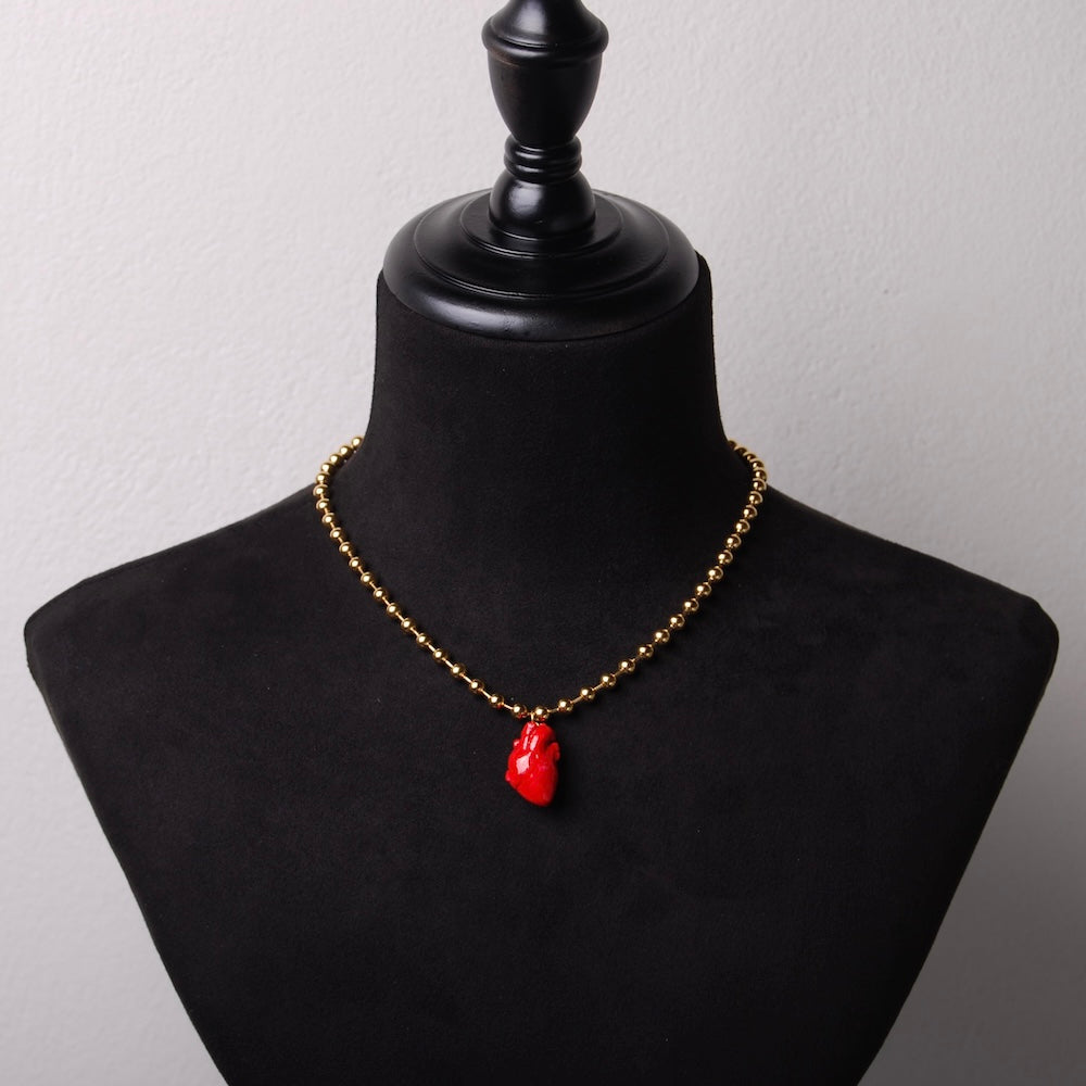 collana con catena a sfere in acciaio placcato oro e pendente cuore anatomico rosso in resina minerale, su busto espositore in velluto nero