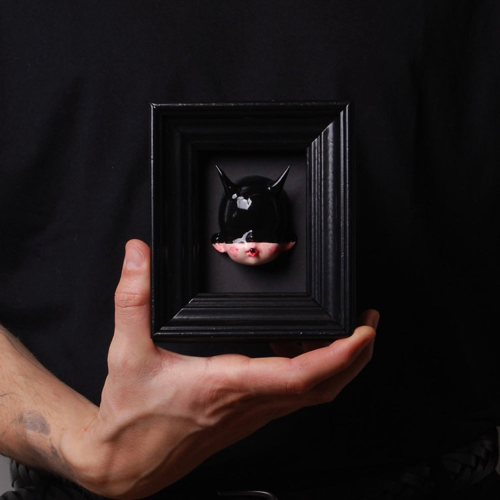 mano che sorregge un quadretto con testa di bambola con maschera nera e corna