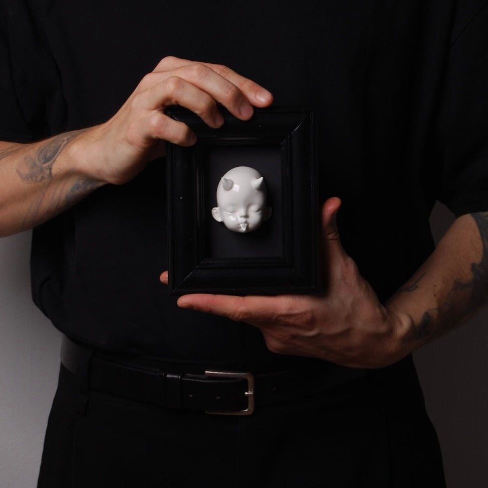 mano che tengono un quadretto testa di bambola bianca con corna dentro ad una cornice nero