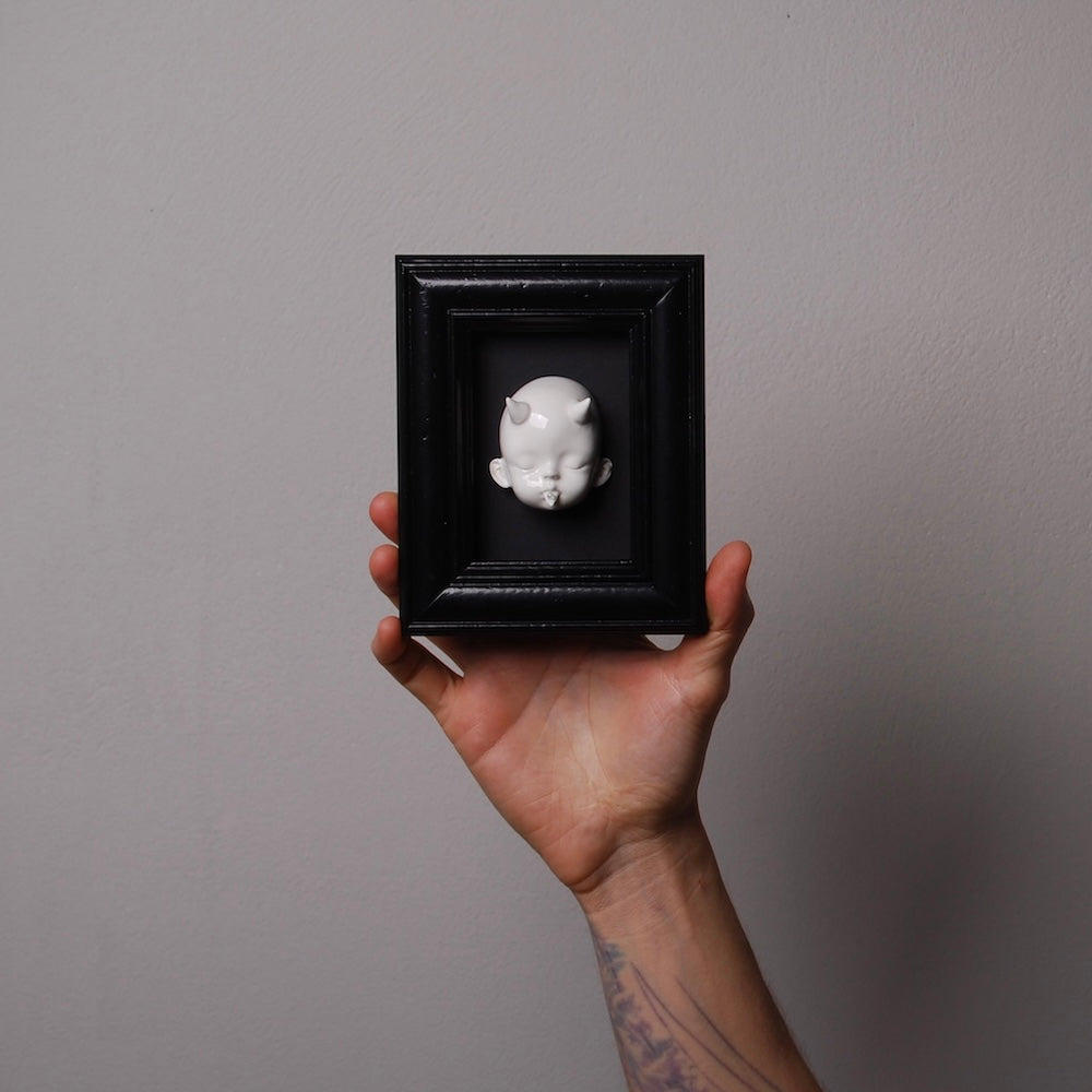 mano che tiene un quadretto nero con testa di bambola bianca con corna