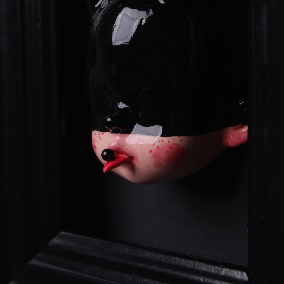 particolare di una testa di bambola all'interno di un quadretto, con maschera nera e lingua fuori dalla bocca con una bolla nera poggiata sopra