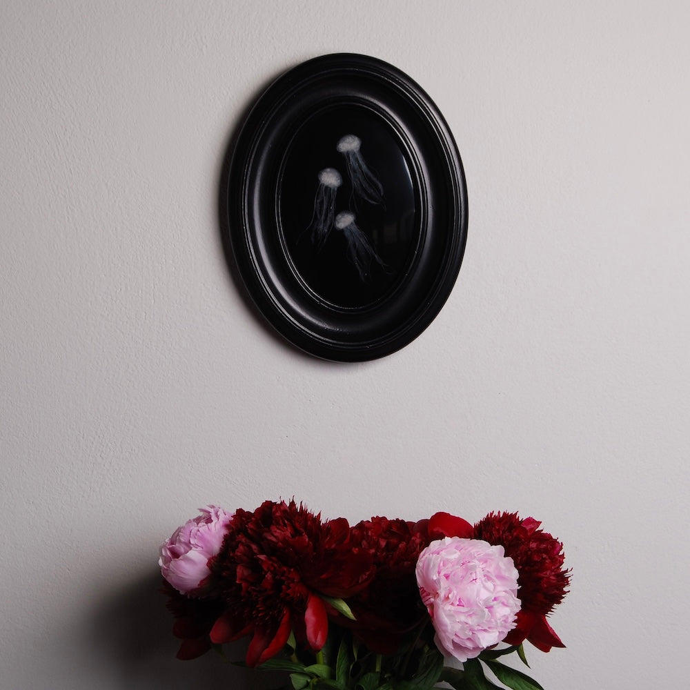 quadretto ovale con meduse sotto resina appeso ad una parete con sotto un vaso di peonie