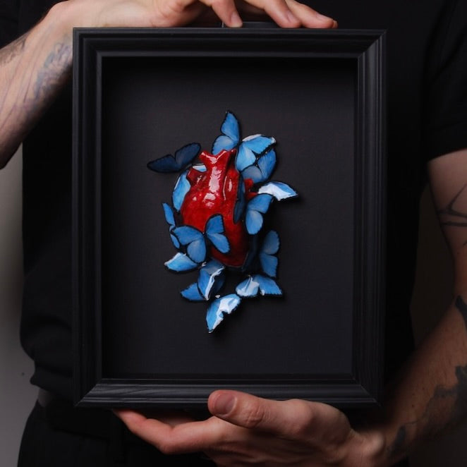 mani che tengono un quadretto con cuore anatomico e sciame di farfalle blu