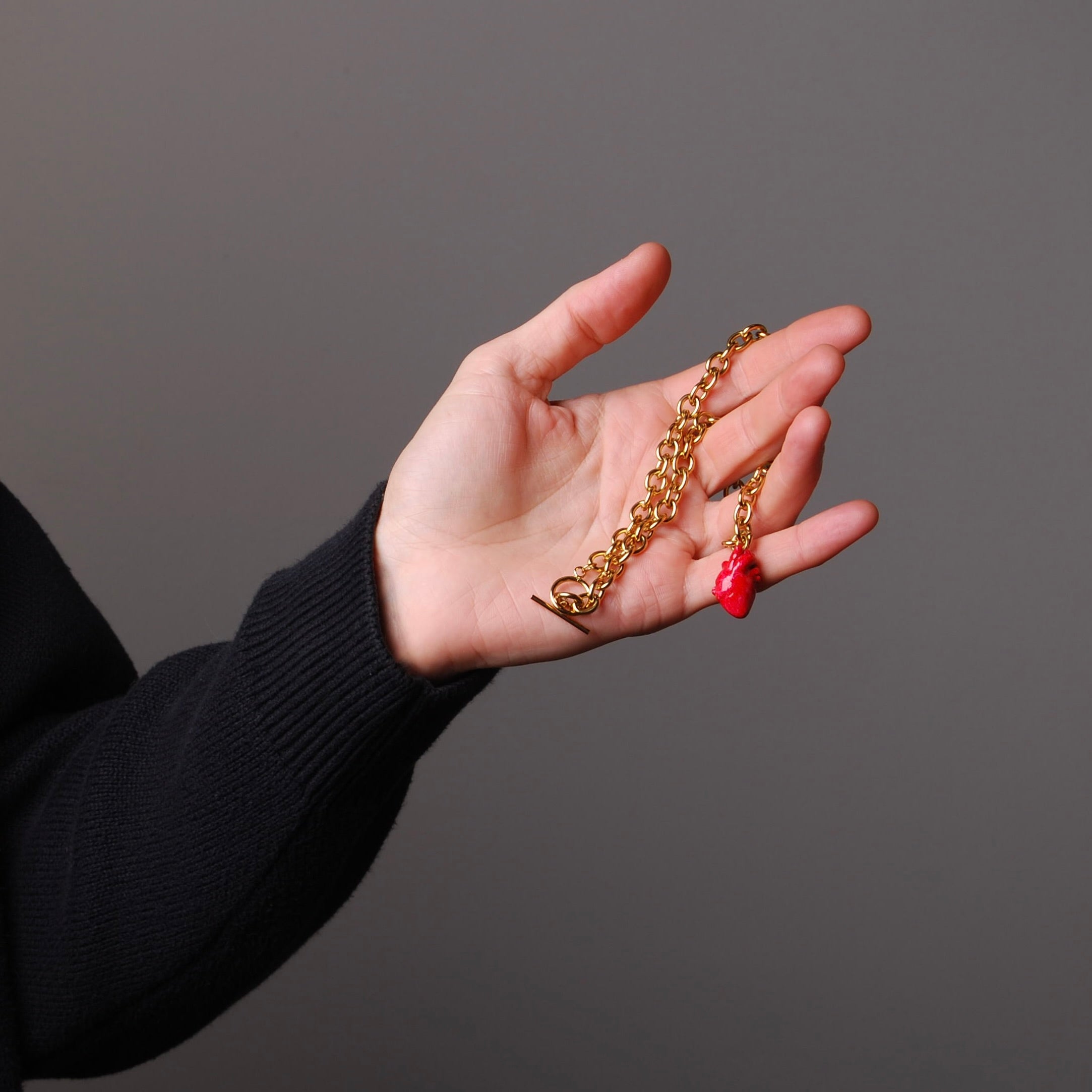 mano che tiene tra le dita una collana a catena spessa con pendente cuore anatomico rosso