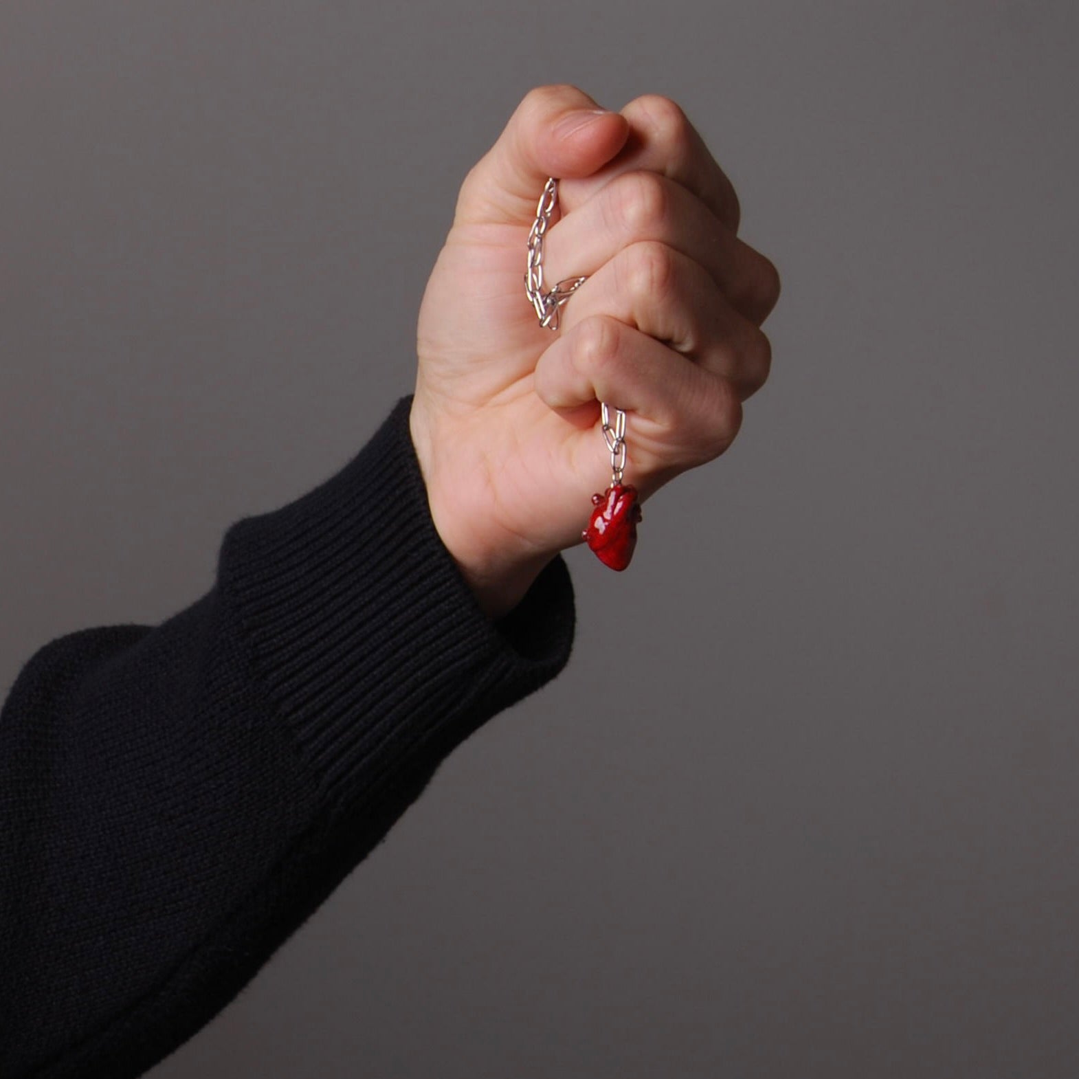 mano che impugna una collana con catena a maglia rettangolare con pendente cuore anatomico rosso