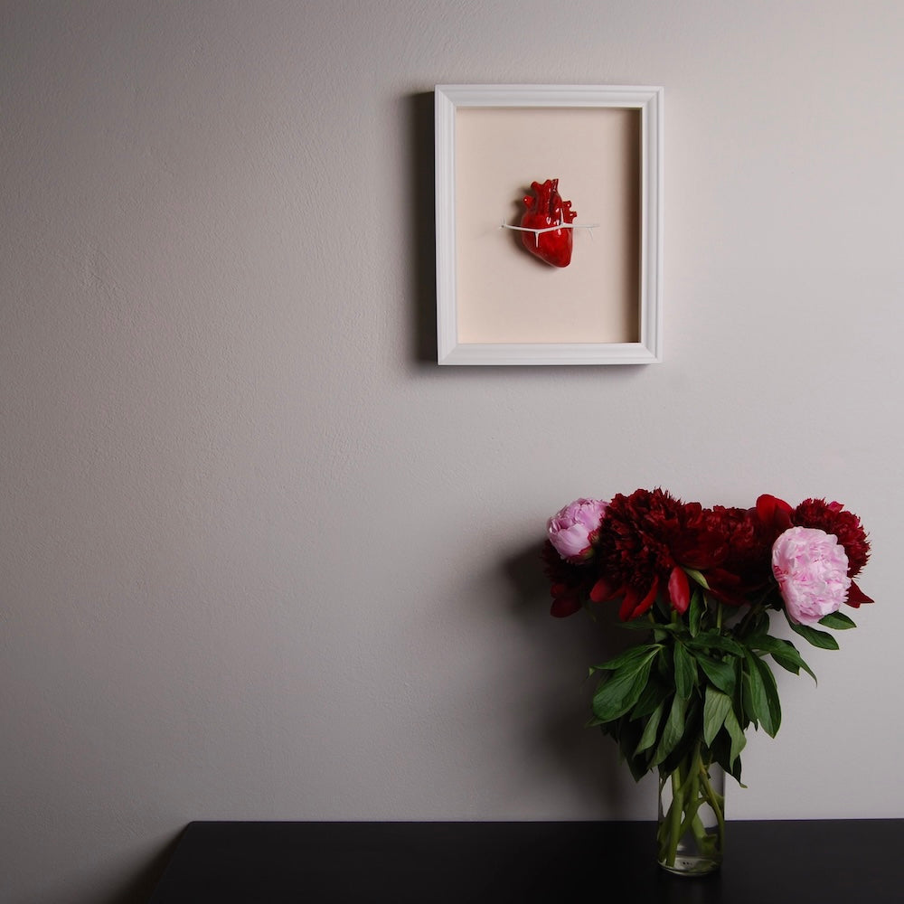 quadretto con cuore anatomico e ramo di spine appeso a una parete, con una scrivania sottostante con sopra un vaso di peonie