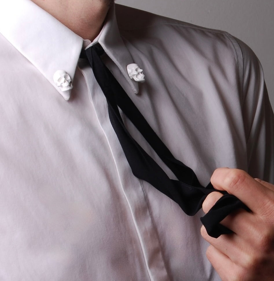 spillette teschio indossate sul collo di una camicia bianca con un nastro nero morbido al centro, tenuto da una mano