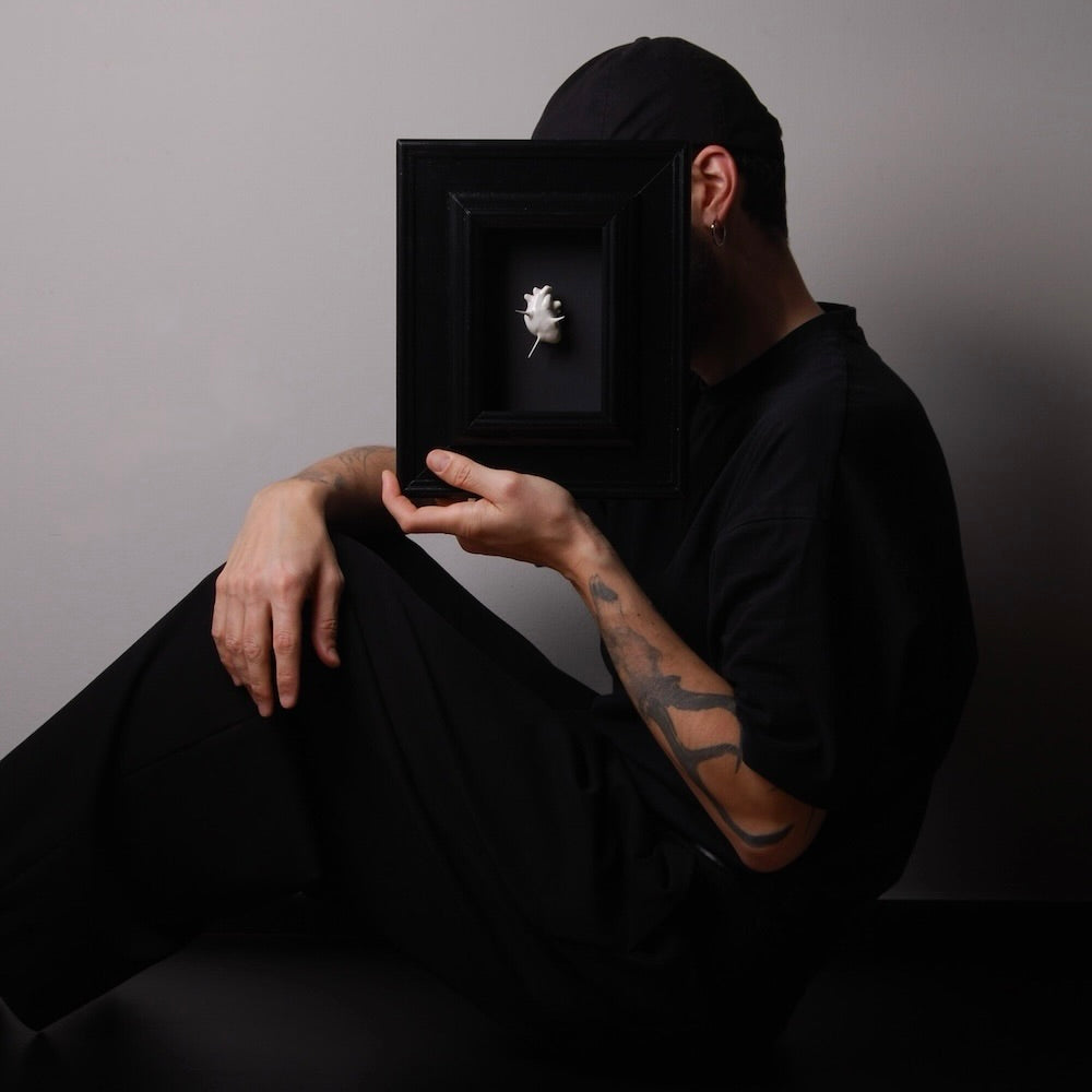 ragazzo seduto per terra che tiene in mano un quadretto con miniatura cuore anatomico bianco spinato