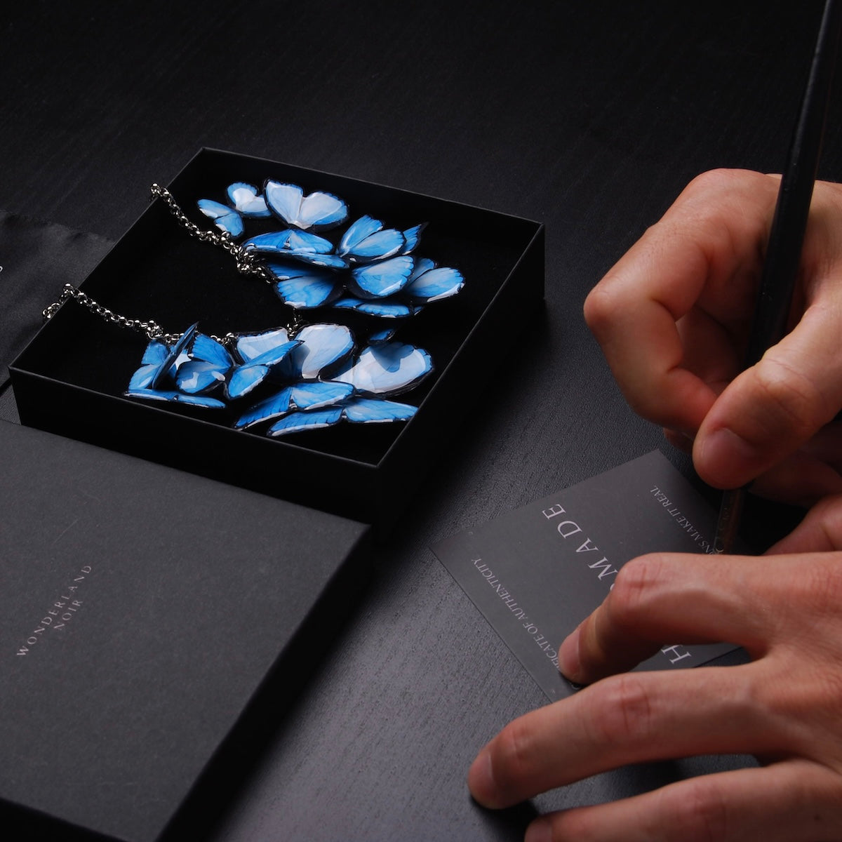 collana sciame di farfalle blu dentro ad una scatolina, con mani che firmano un certificato di autenticità