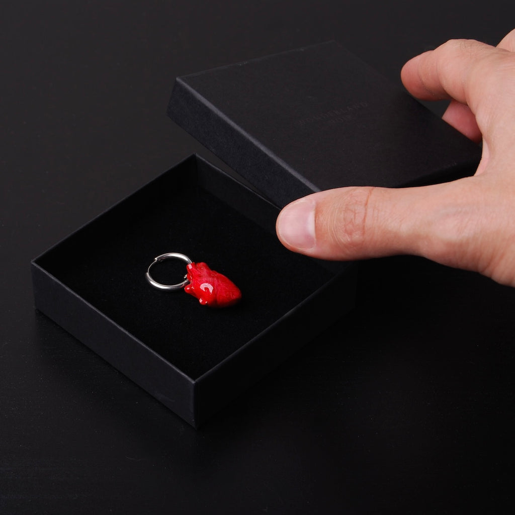 mano che chiude una scatolina con un orecchino singolo con pendente cuore anatomico rosso