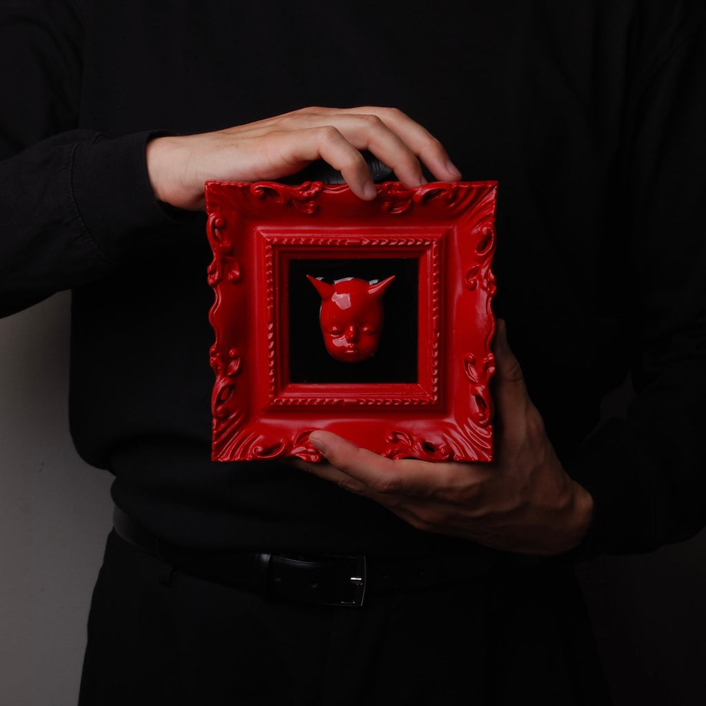 mani che tengono un quadretto barocco rosso con testa di diavoletto rosso