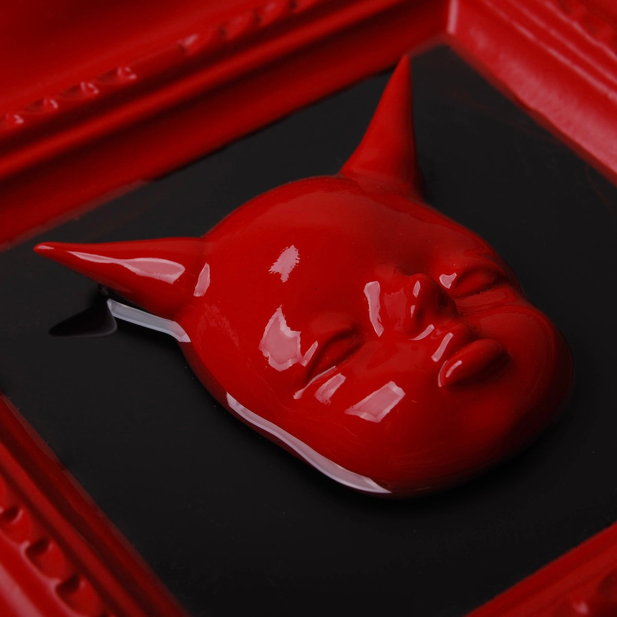 dettaglio del volto di un diavoletto rosso con corna lunghe, al centro di una cornice barocca rossa