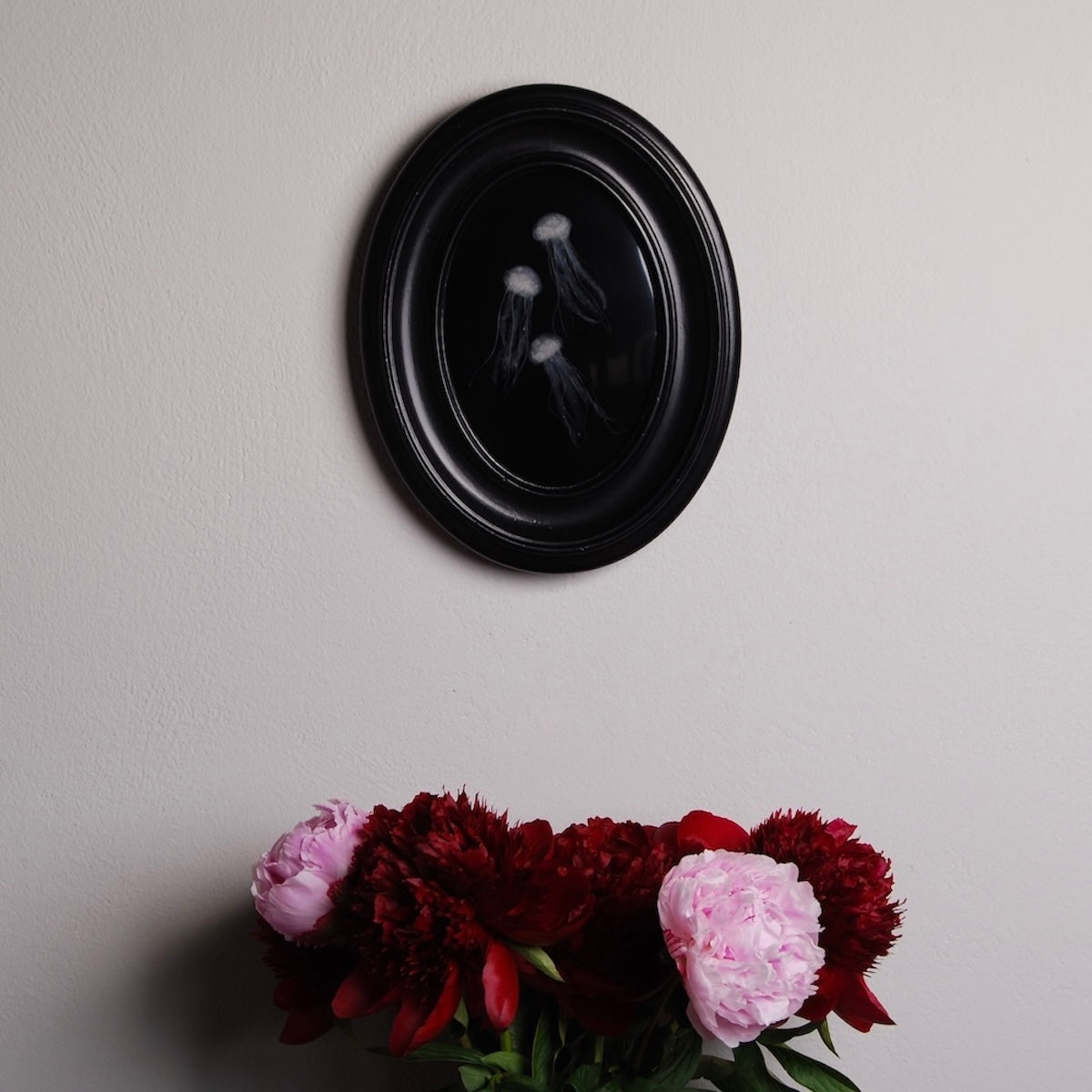 quadro ovale con meduse marine appeso al muro, con sotto un vaso di peonie