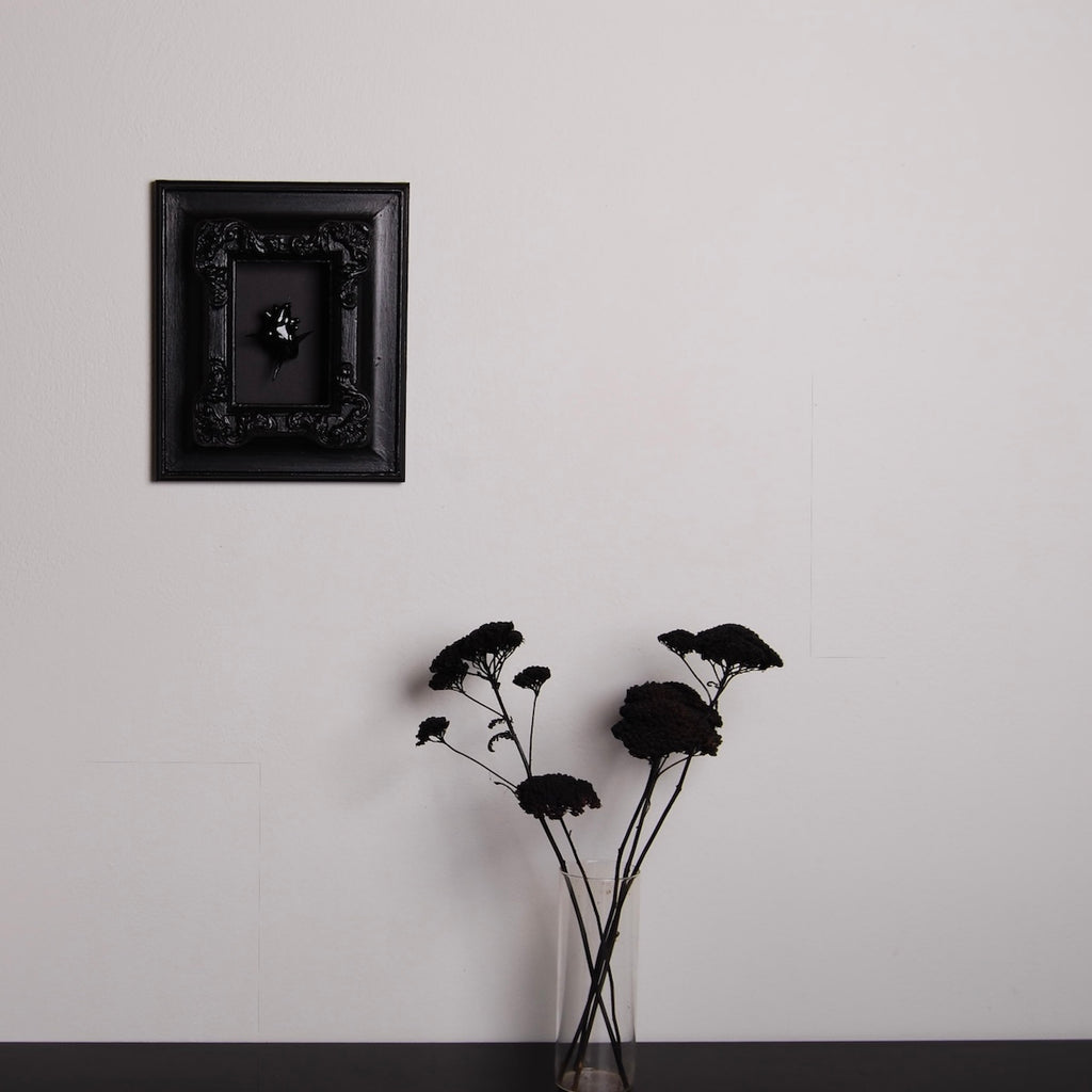un quadro con cornice antica con cuore anatomico spinato nero appeso ad una parete, con sottostante un vaso di fiori neri su piano nero