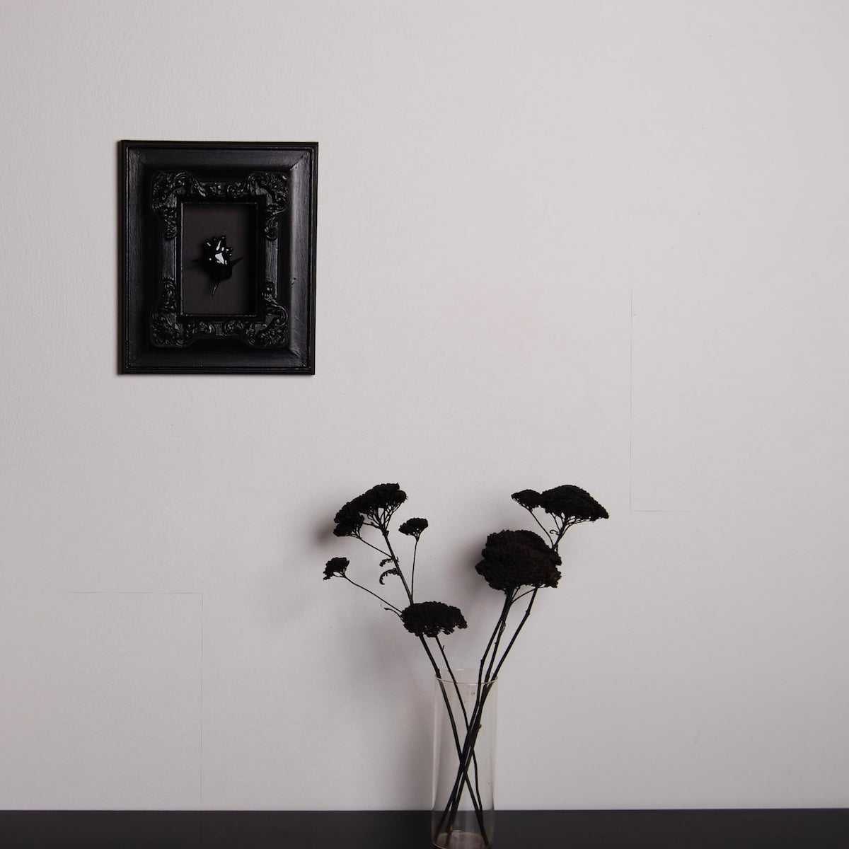 un quadro con cornice antica con cuore anatomico spinato nero appeso ad una parete, con sottostante un vaso di fiori neri su piano nero