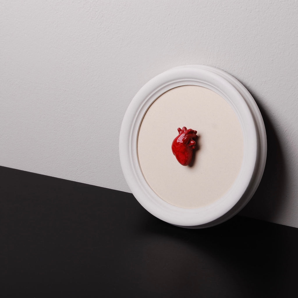 quadretto rotondo con cornice bianca e cuore anatomico rosso poggiato al muro sopra un ripiano nero