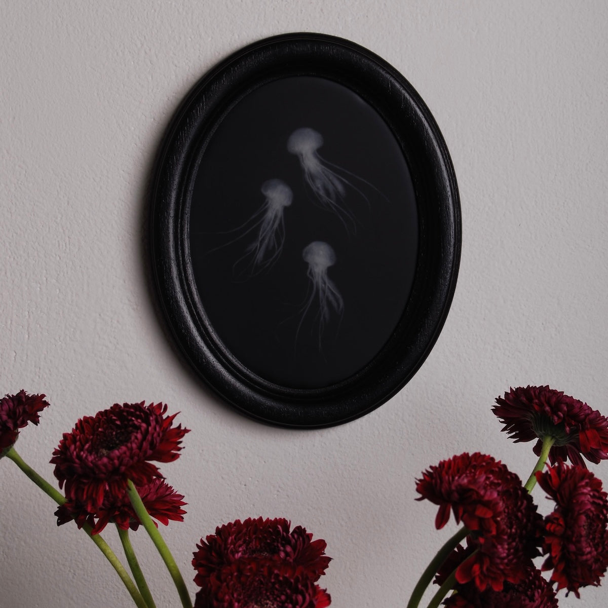 quadro ovale nero con 3 sculture meduse marine sospeso sopra gerbere amaranto